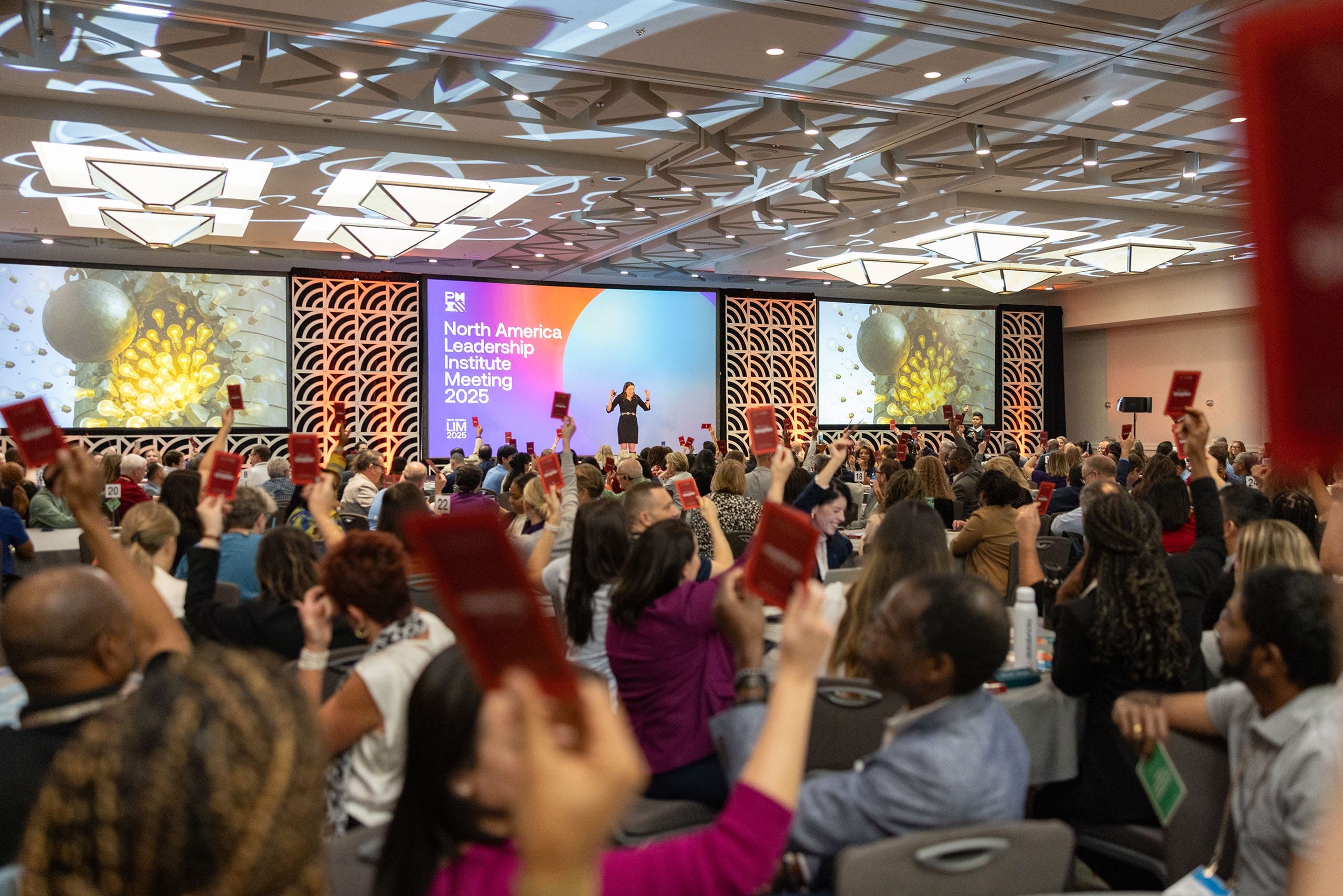 A keynote speaker stands on a large conference stage addressing a packed audience, with attendees raising red cards during an interactive session at the North America Leadership Institute Meeting.